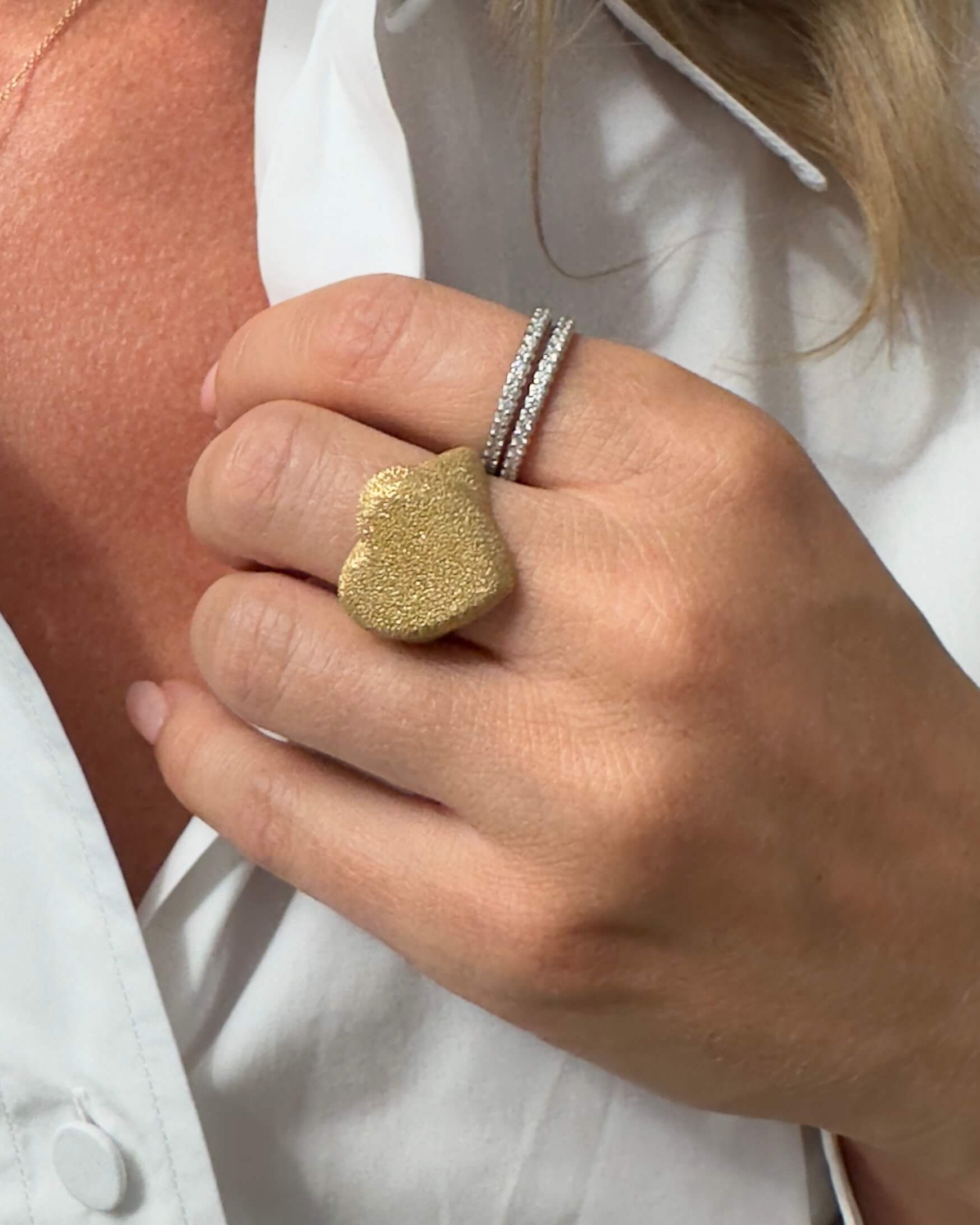 Close-up of a hand wearing a gold textured heart ring with a white shirt background.