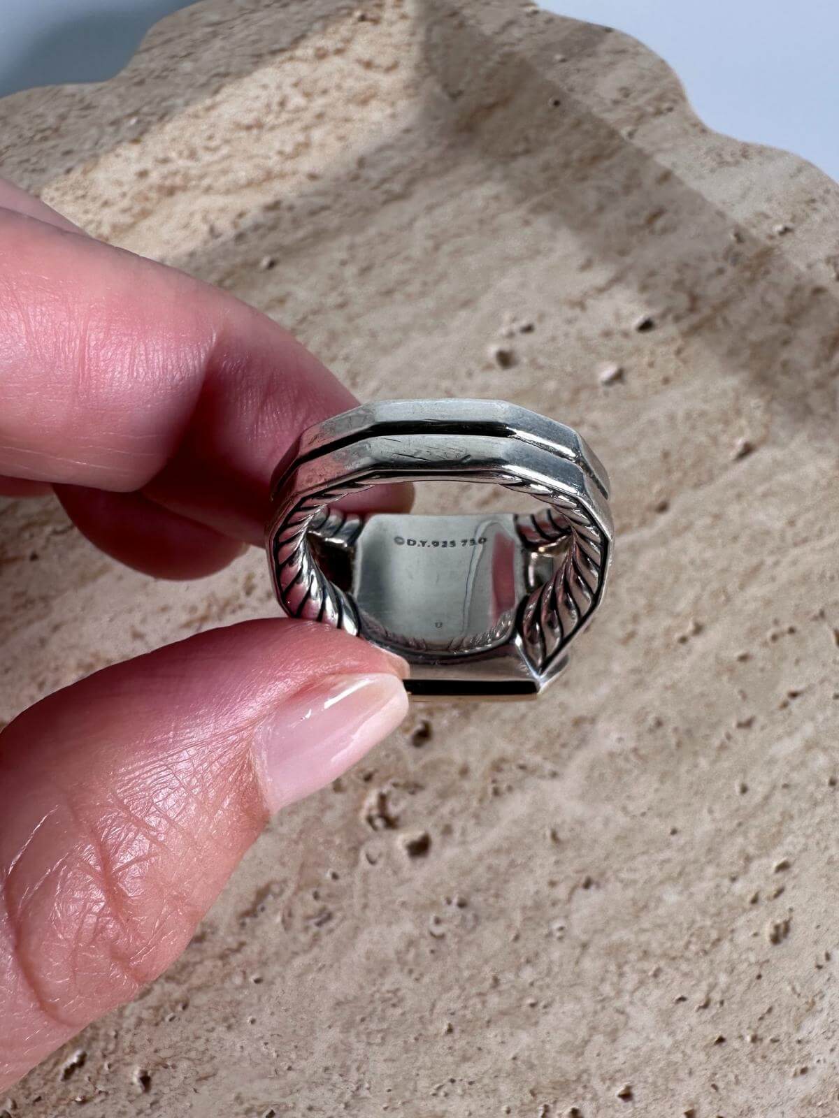 DY Silver ring held between fingers on a textured surface showing signature on back of ring
