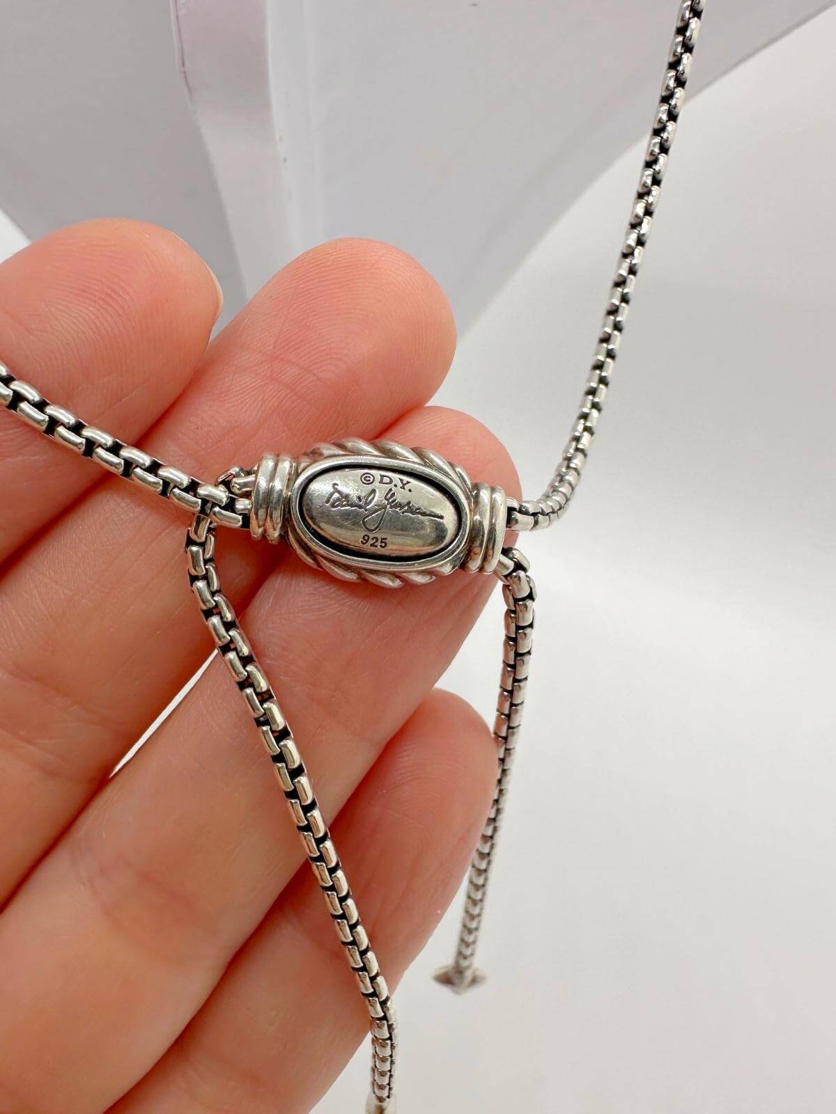 Signature of DY on silver necklace held by hand on white background