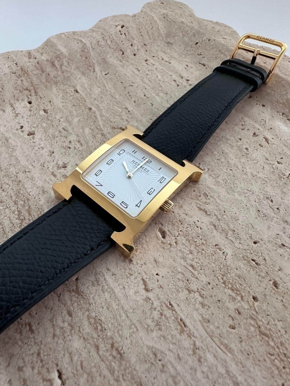 Hermes H Heure Gold-framed watch with black strap on a textured surface