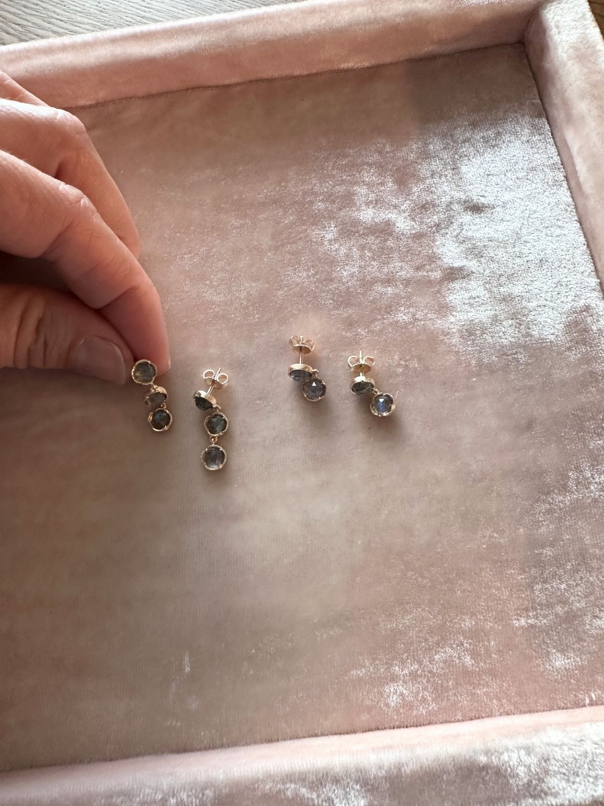 Louise Row Studio labradorite drop 18K gold earrings shown on a pink surface with a hand holding one earring.
