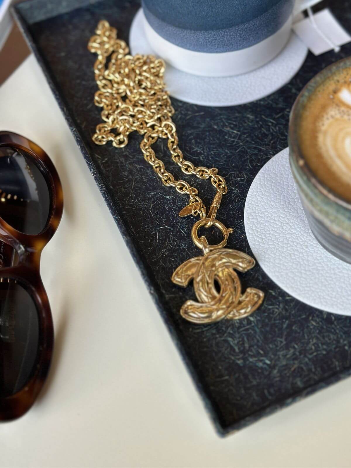 Gold necklace with a Chanel pendant on a table next to sunglasses and a coffee cup.