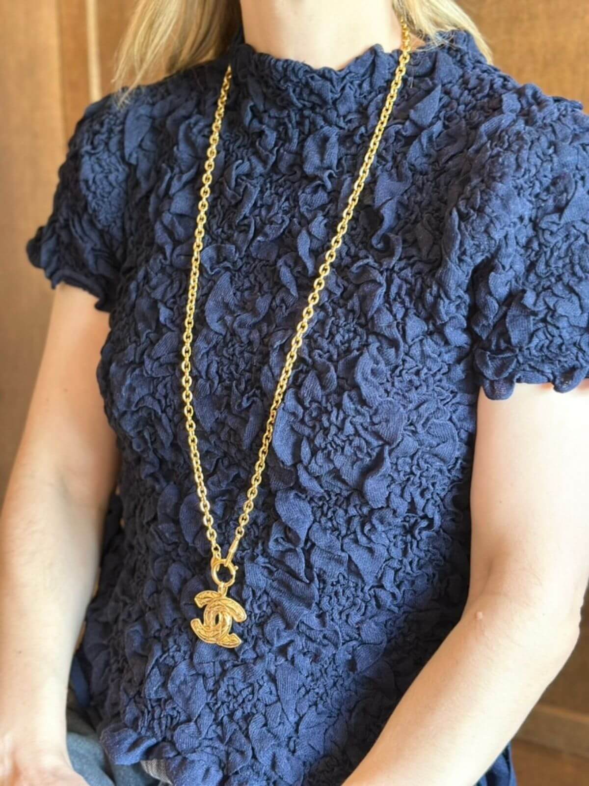 Person wearing a navy lace top with a gold chain necklace featuring a logo pendant.