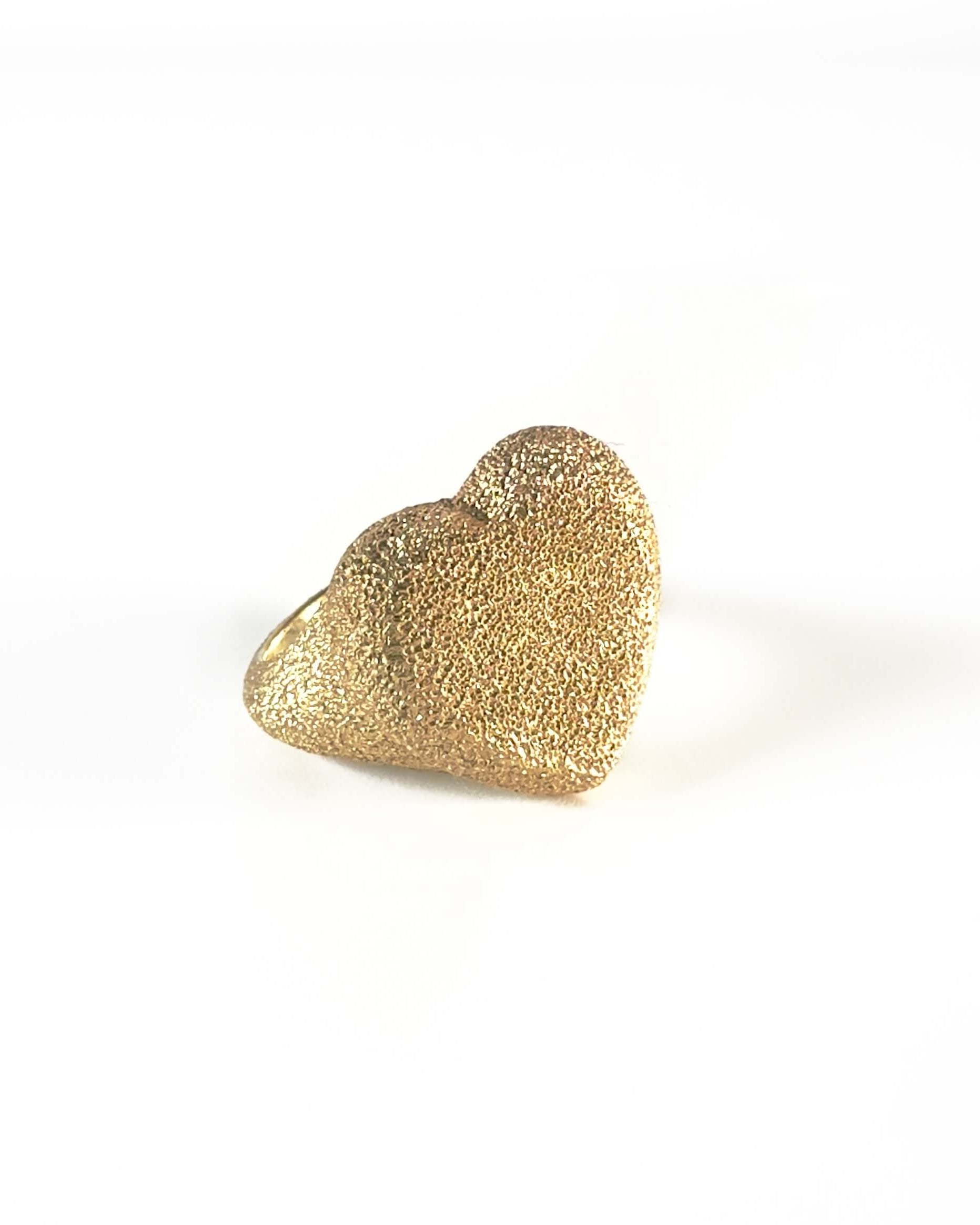 Carolina Bucci gold ring in the shape of a heart , facing the left with a white background. The ring has a sparkle due to the texture. 