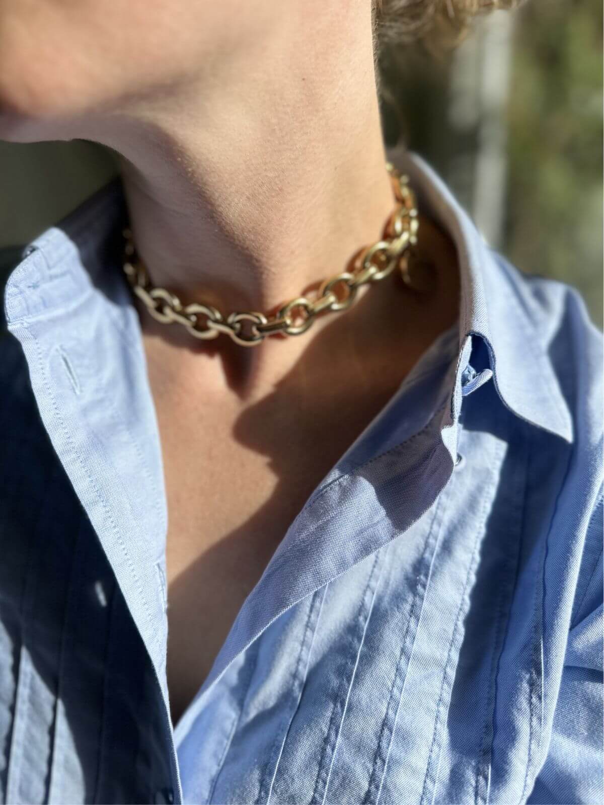 Close-up of a person wearing a gold chain necklace with a blurred outdoor background