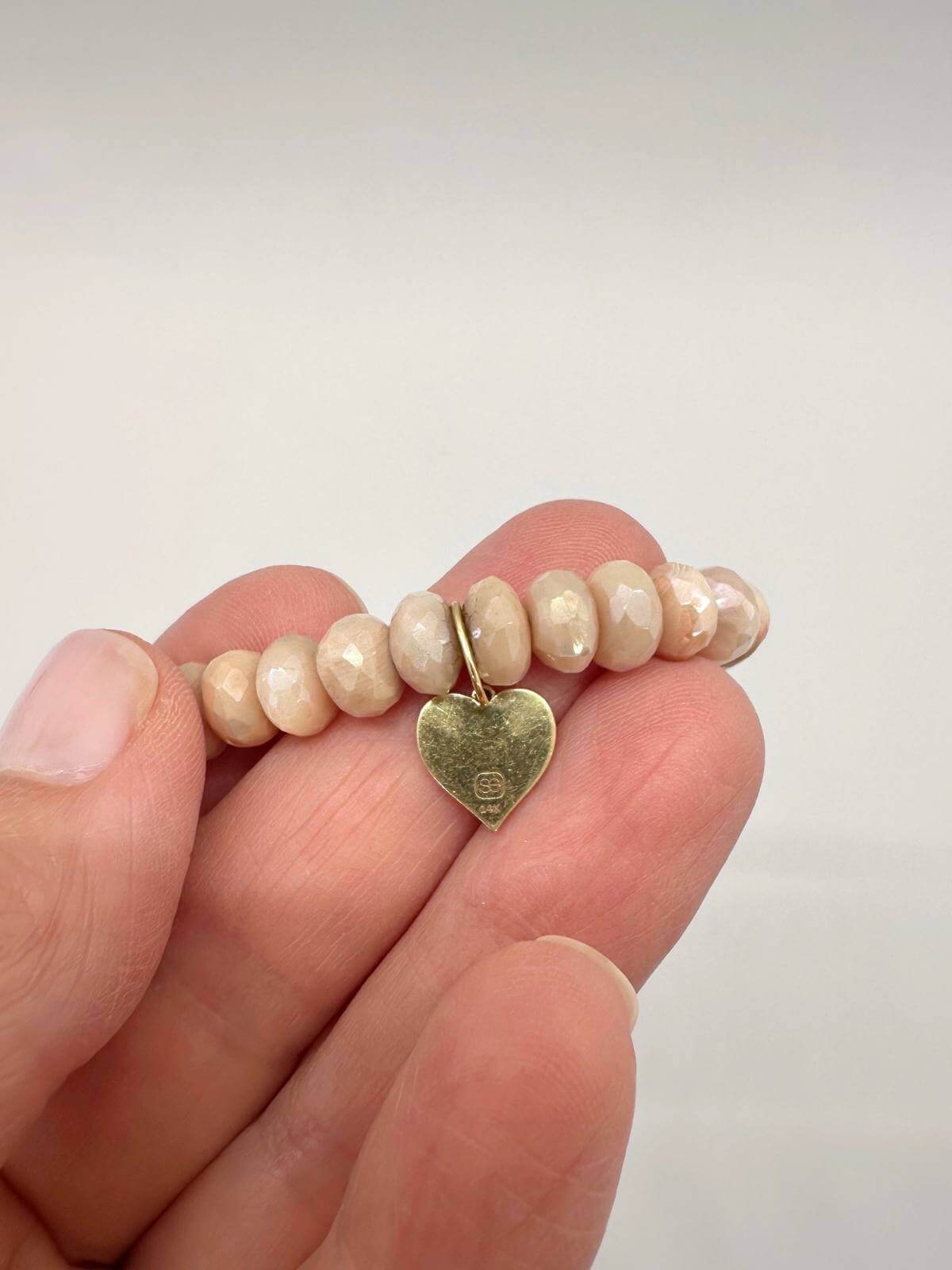 Hand holding a beaded bracelet with a gold heart charm against a plain background