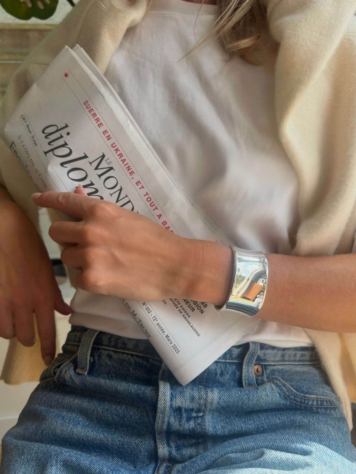 Person holding a newspaper wearing a silver Tiffany bracelet with 'Le Monde' visible, wearing a beige cardigan and blue jeans.