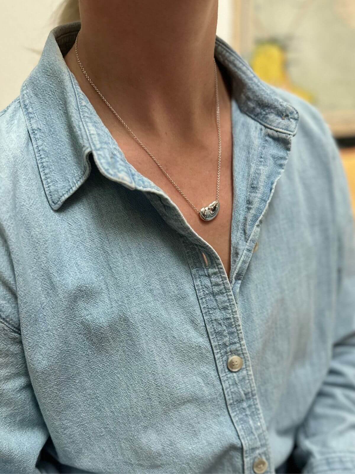 Person wearing a light blue denim shirt with a necklace featuring a bean pendant.