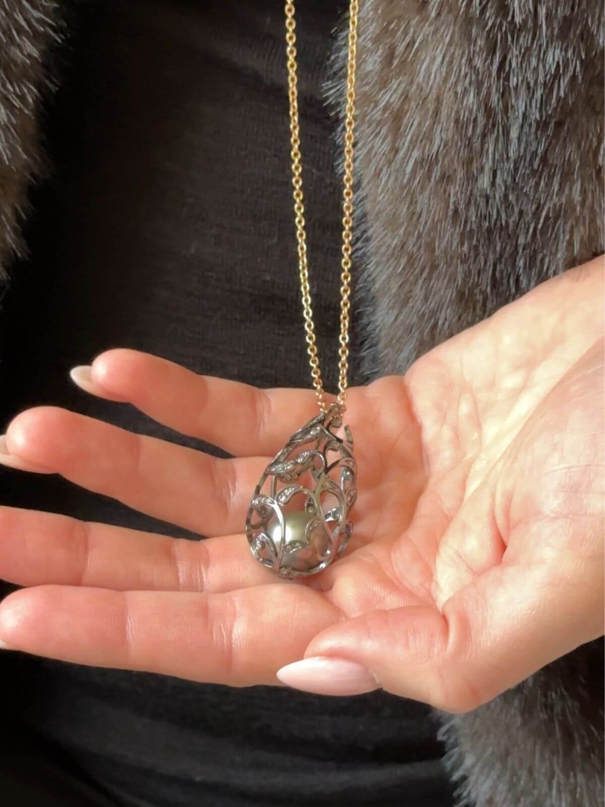 Louise Row Studio featuring Utopia Bolero pearl and diamond necklace on model with fur coat detail of Hand holding a silver teardrop-shaped pendant on a gold chain against a dark background.
