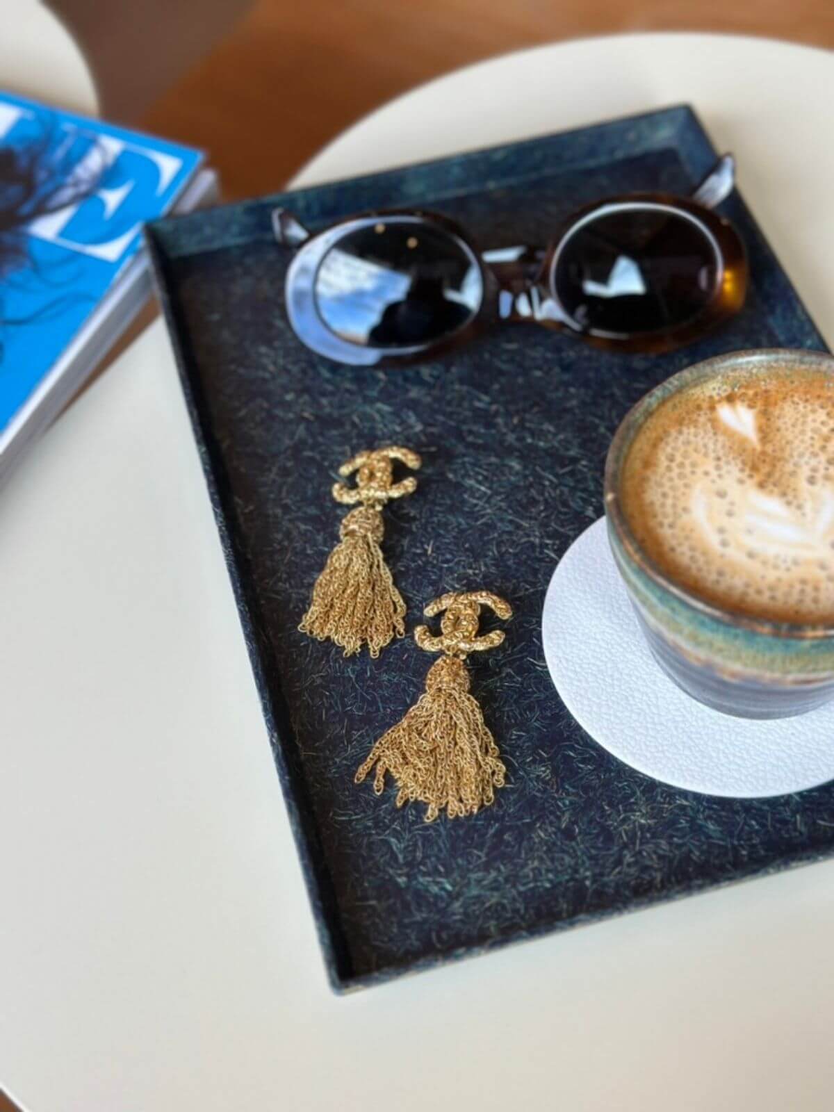 Gold tassel earrings on a dark surface with sunglasses and a cappuccino in the background.